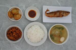 A meal consisting of a plate of fried fish, a bowl of white rice, a bowl of vegetable soup with meatballs, a small plate of red sauce and others, all on a gray marble surface. photo