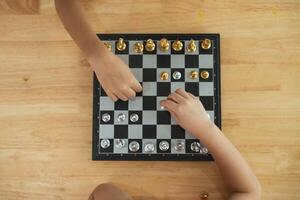 Two children playing a game of chess on a wooden table. One of the children is holding a piece in his hand photo