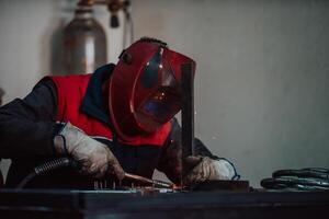Profesional welder in protective uniform and mask welding metal pipe on the industrial table with other workers behind in the industrial workshop. photo