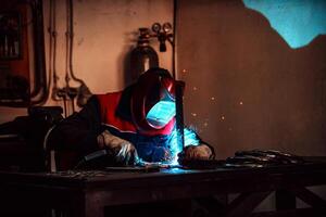 Profesional welder in protective uniform and mask welding metal pipe on the industrial table with other workers behind in the industrial workshop. photo