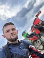 A professional motorcyclist resting on a forest road after an adventurous ride and taking a selfie for social networks photo