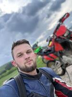 A professional motorcyclist resting on a forest road after an adventurous ride and taking a selfie for social networks photo