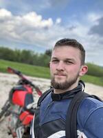 A professional motorcyclist resting on a forest road after an adventurous ride and taking a selfie for social networks photo