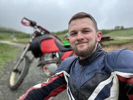 A professional motorcyclist resting on a forest road after an adventurous ride and taking a selfie for social networks photo