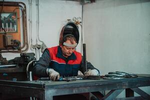Profesional welder in protective uniform and mask welding metal pipe on the industrial table with other workers behind in the industrial workshop. photo