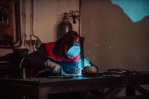 Profesional welder in protective uniform and mask welding metal pipe on the industrial table with other workers behind in the industrial workshop. photo