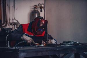 Profesional welder in protective uniform and mask welding metal pipe on the industrial table with other workers behind in the industrial workshop. photo