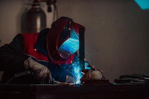 Profesional welder in protective uniform and mask welding metal pipe on the industrial table with other workers behind in the industrial workshop. photo
