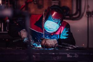 Profesional welder in protective uniform and mask welding metal pipe on the industrial table with other workers behind in the industrial workshop. photo