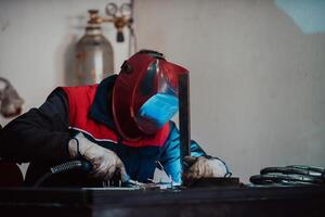 Profesional welder in protective uniform and mask welding metal pipe on the industrial table with other workers behind in the industrial workshop. photo