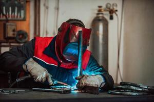 Profesional welder in protective uniform and mask welding metal pipe on the industrial table with other workers behind in the industrial workshop. photo