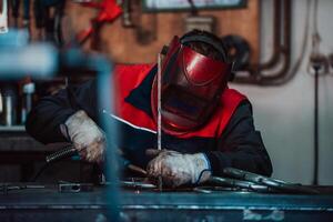 Profesional welder in protective uniform and mask welding metal pipe on the industrial table with other workers behind in the industrial workshop. photo