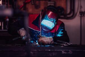 Profesional welder in protective uniform and mask welding metal pipe on the industrial table with other workers behind in the industrial workshop. photo