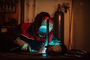 Profesional welder in protective uniform and mask welding metal pipe on the industrial table with other workers behind in the industrial workshop. photo