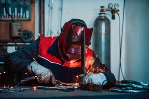 Profesional welder in protective uniform and mask welding metal pipe on the industrial table with other workers behind in the industrial workshop. photo