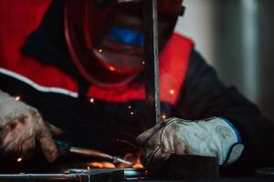 Close up welder in protective uniform and mask welding metal pipe on the industrial table with other workers behind in the industrial workshop. photo