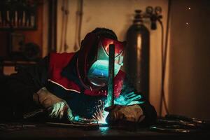 Profesional welder in protective uniform and mask welding metal pipe on the industrial table with other workers behind in the industrial workshop. photo