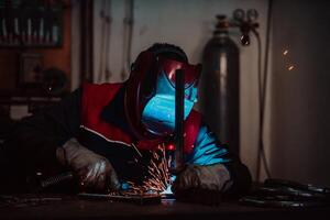 Profesional welder in protective uniform and mask welding metal pipe on the industrial table with other workers behind in the industrial workshop. photo