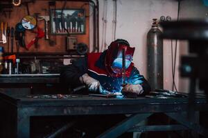 Close up welder in protective uniform and mask welding metal pipe on the industrial table with other workers behind in the industrial workshop. photo