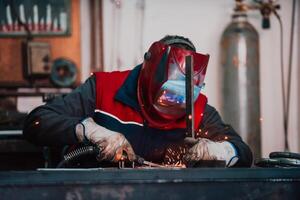 Profesional welder in protective uniform and mask welding metal pipe on the industrial table with other workers behind in the industrial workshop. photo