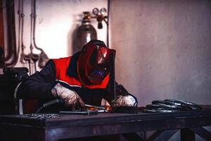 Profesional welder in protective uniform and mask welding metal pipe on the industrial table with other workers behind in the industrial workshop. photo