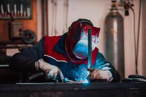 Profesional welder in protective uniform and mask welding metal pipe on the industrial table with other workers behind in the industrial workshop. photo