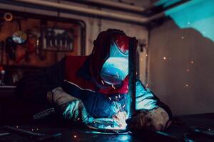 Profesional welder in protective uniform and mask welding metal pipe on the industrial table with other workers behind in the industrial workshop. photo