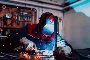 Profesional welder in protective uniform and mask welding metal pipe on the industrial table with other workers behind in the industrial workshop. photo