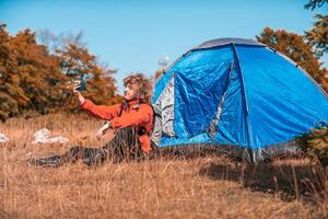 A man with a tent enjoys a sunny day whileusing a smartphone and camping in the mountains photo