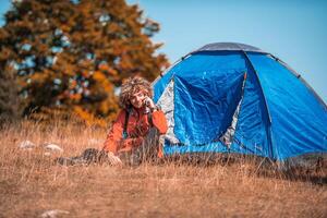 A man with a tent enjoys a sunny day whileusing a smartphone and camping in the mountains photo