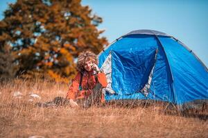 A man with a tent enjoys a sunny day whileusing a smartphone and camping in the mountains photo