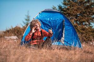 A man with a tent enjoys a sunny day whileusing a smartphone and camping in the mountains photo