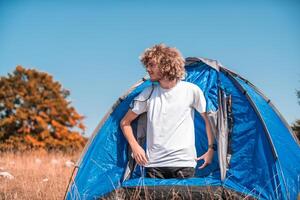 A man with a tent enjoys a sunny day whileusing a smartphone and camping in the mountains photo