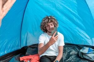 A man with a tent enjoys a sunny day whileusing a smartphone and camping in the mountains photo