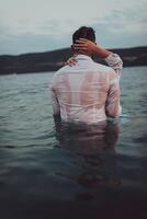 A symbolic and emotionally connected couple tenderly embraces against the backdrop of a serene sea, bathed in the warm hues of a breathtaking sunset photo