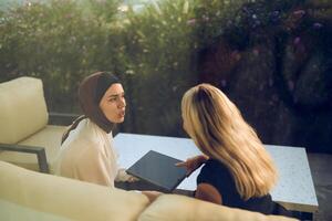 A hijab-wearing woman, a successful real estate business owner, gracefully sells a house to a blonde customer, both sitting in front of the property. photo