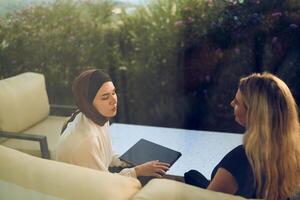 A hijab-wearing woman, a successful real estate business owner, gracefully sells a house to a blonde customer, both sitting in front of the property. photo