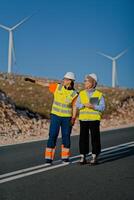 A engineer with a tablet and her colleague inspect wind turbines in a field, demonstrating teamwork and dedication to renewable energy projects photo