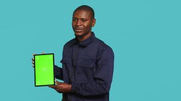 Portrait of smiling security guard at work showcasing isolated screen tablet while overseeing premises. Joyful private law enforcement worker presenting chroma key device, studio backdrop, camera A video