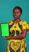 Vertical Feminine person in native clothing holds a tablet with mockup screen, creating an advertisement against blue background in studio. Young woman smiling and presenting copy space. Camera B. video