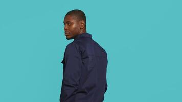 Security guard turning around after hearing suspect sounds during surveillance shift, ensuring protection. Portrait of man in uniform standing isolated with back against studio background, camera A video