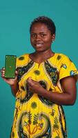Vertical Young confident woman presenting a green screen on smartphone, working in studio on an advertisement. Smiling natural girl holds a phone with isolated chroma key display. Camera B. video