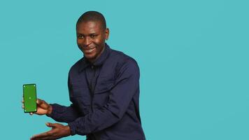 Portrait of upbeat security staff member showing green screen smartphone. Cheerful patrolling agent maintains secure environment, showcasing mockup phone, studio background, camera A video
