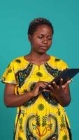 Vertical Natural cute girl browsing internet on a tablet against blue background, navigating on online web pages by using a portable device. Woman posing with a gadget and traditional outfit. Camera B. video