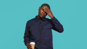 Tired security guard drinking coffee cup to stay awake, being tasked with preventing criminals. Portrait of weary bodyguard sipping caffeinated beverage during night shift, studio background, camera A video