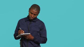 Portrait of security guard using notepad to take notes of suspicious activity while overseeing premises. Private law enforcement worker documenting security protocols, studio backdrop, camera A video
