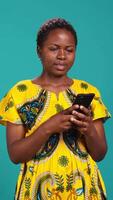 Vertical Native girl in ethnic outfit browsing internet webpages in studio, using her smartphone to check texts and social media platforms. Young person standing against blue background. Camera B. video