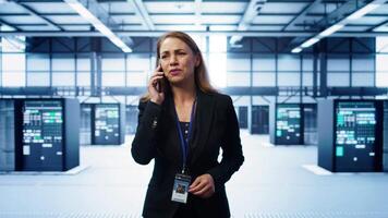 Software engineer talking on smartphone in data center, coordinating server maintenance tasks. Woman surrounded by server racks and high tech equipment, speaking during telephone call, camera B video
