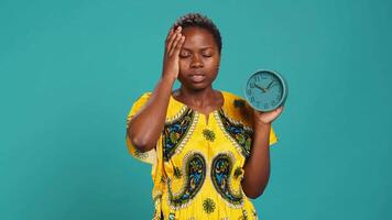 Young woman checking the time on a clock and running late, trying to hurry and stop the delay against blue background. Busy person missing the alarm, having problems with punctuality. Camera A. video