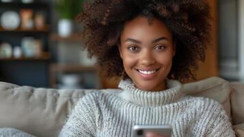 un mujer con Rizado pelo sonrisas calurosamente mientras sentado en un sofá y mirando a su teléfono foto